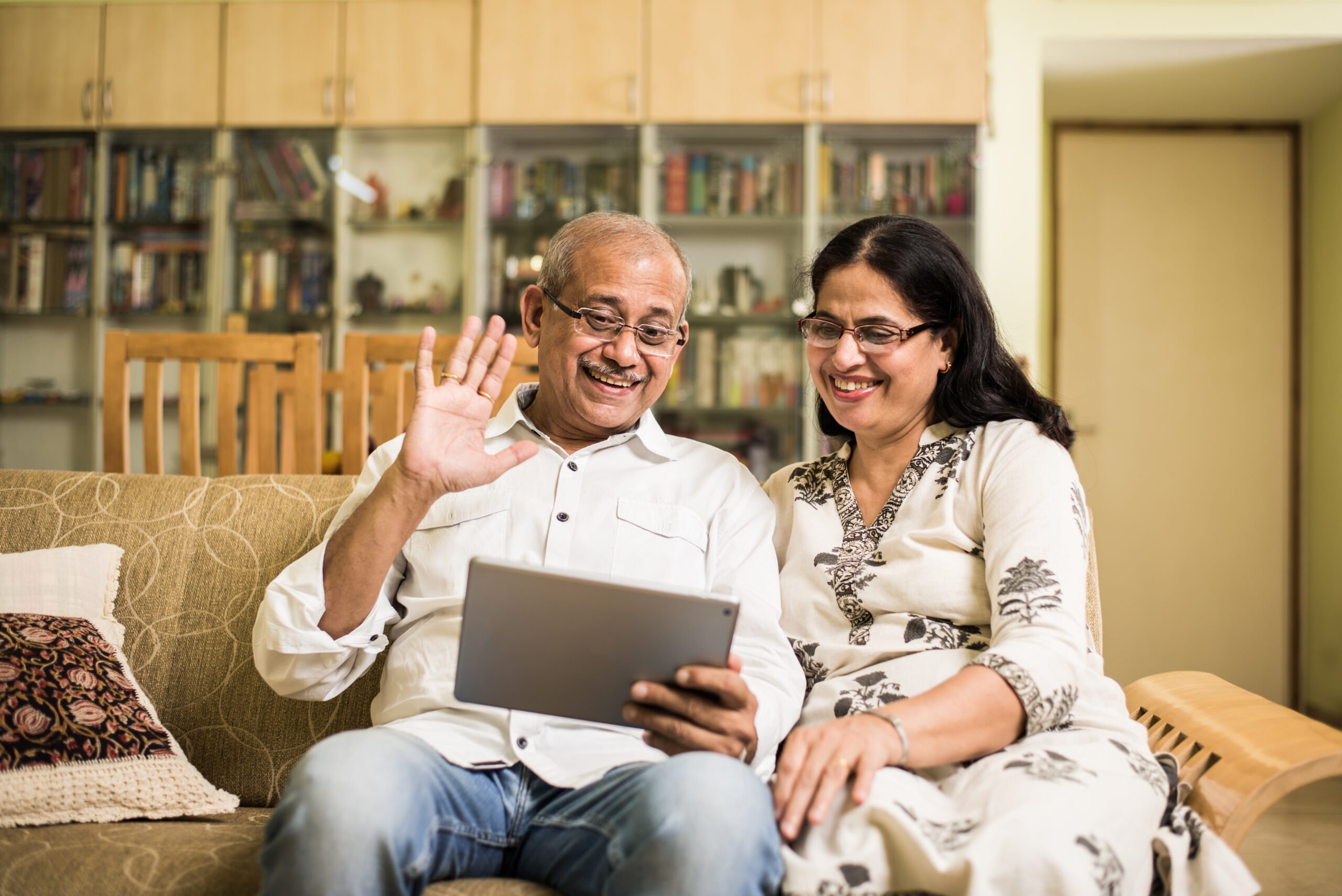 Two Indian people sat on a couch. The man on the left of the image is wearing glasses and is waving at a tablet screen, the woman on the right is wearing glasses and is smiling. They are in a home environment. 