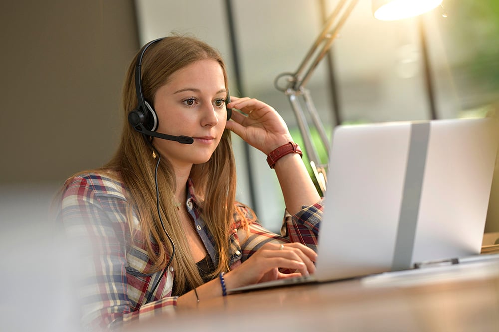 Young Caucasian woman wearing a headset and using a laptop at a desk. She is wearing a checked shirt.