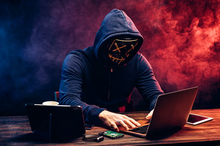 hacker man typing on laptop, hacking computer system. male in mask and pullover. unrecognizable incognito male sit in hood and try to breach the security of laptop system. neon smoke in background