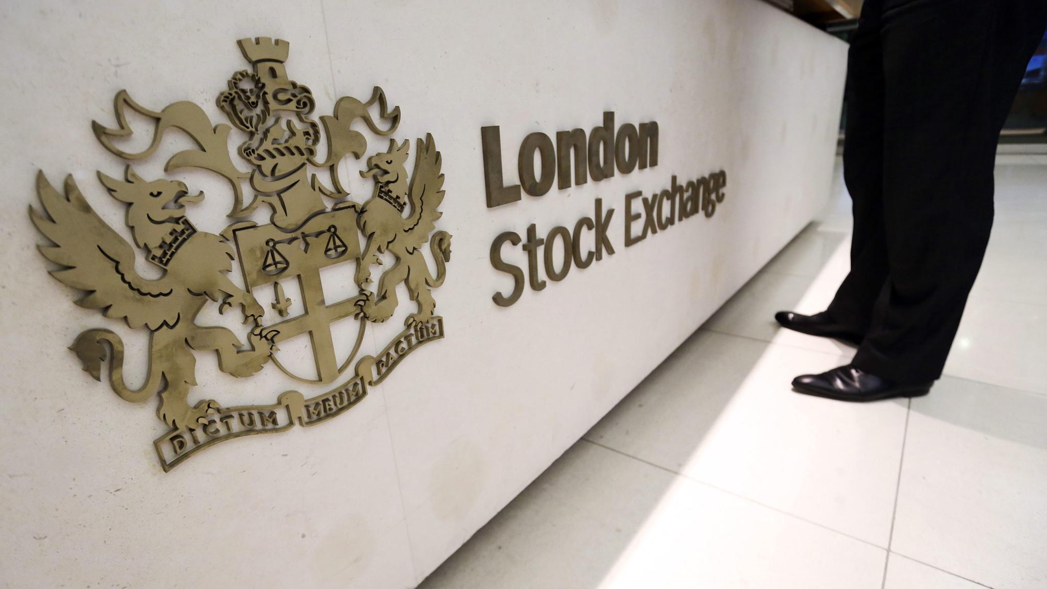 A man in a black suit and black shoes stands behind a board displaying the logo of the London Stock Exchange Group PLC.