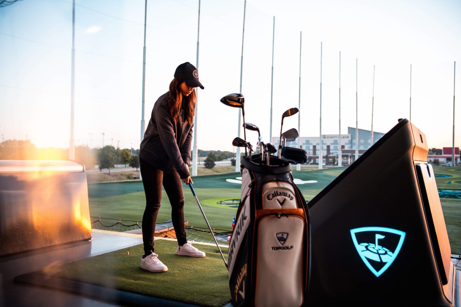A girl in black hitting ball with a golf stick, the golf equipment bag of Callaway Golf Co. is placed on a side.