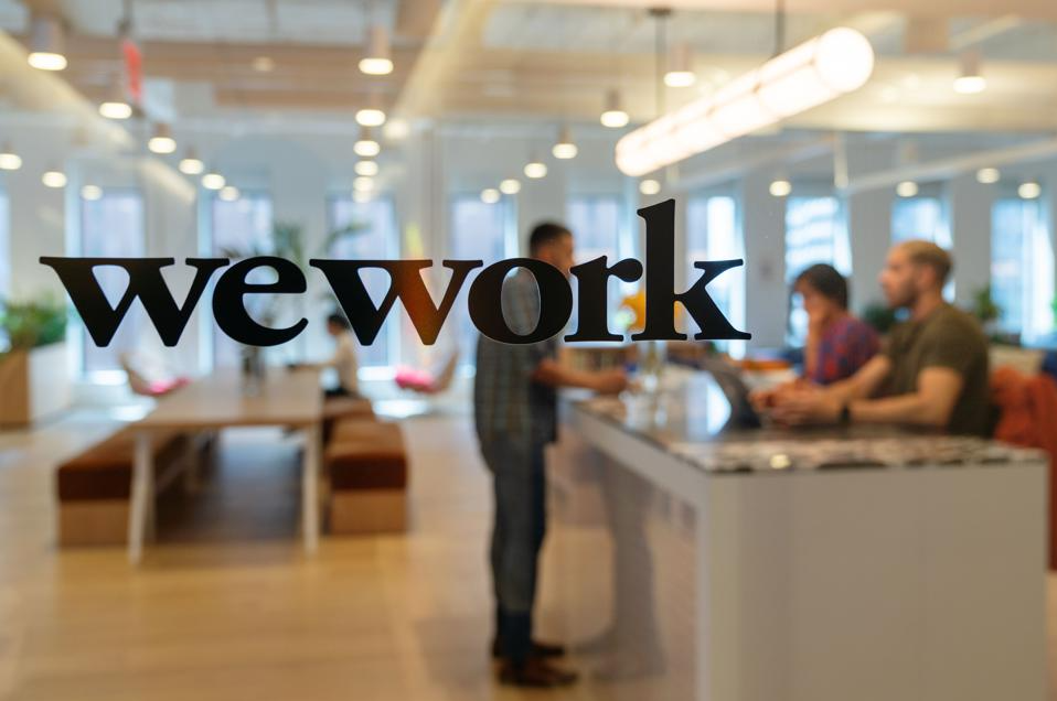 Wework Inc's logo is placed on a photo with a blurry background of an office counter with multiple employees sitting and a person standing