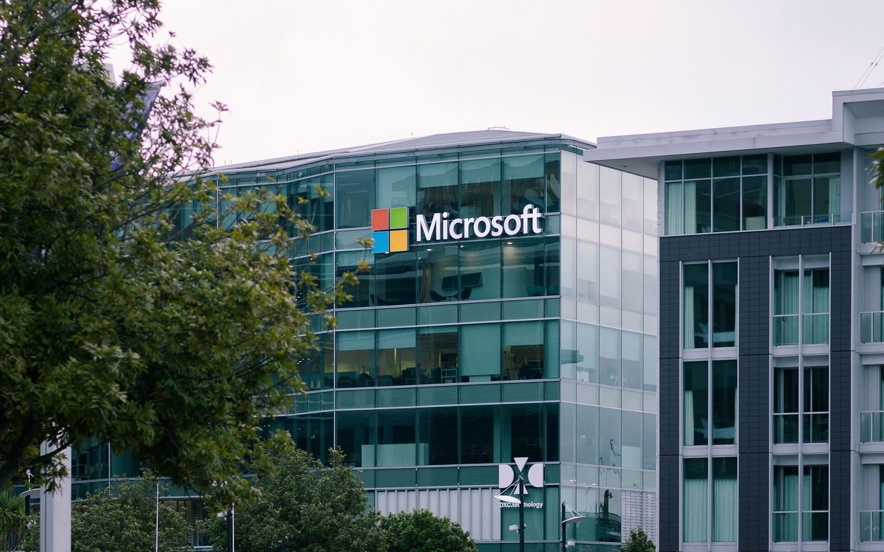 The Microsoft Corporation logo is mounted on a blue and white mirror building with a tree planted behind it.
