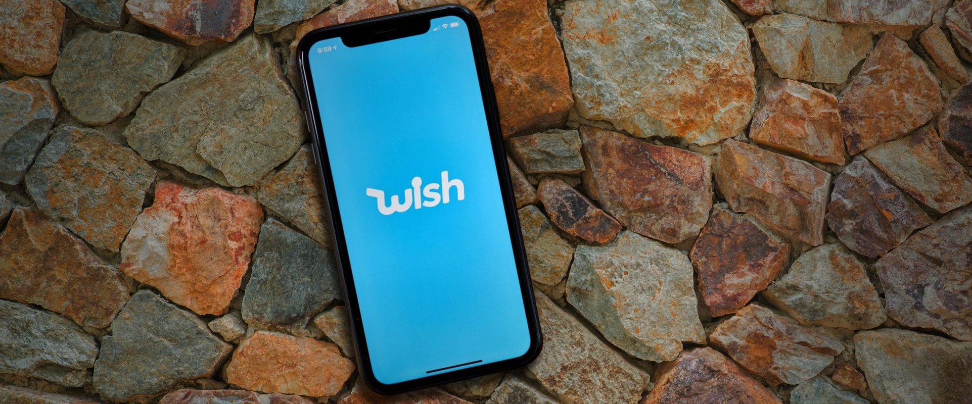 A mobile phone with a sky blue wallpaper and the WISH logo, placed atop stones.