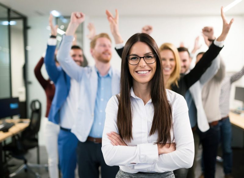 Smiling woman in an office with arms folded with colleagues celebrating behind her