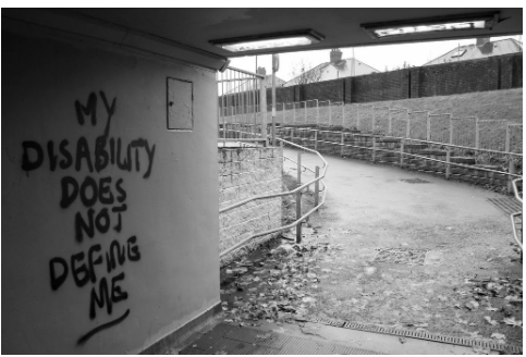  A black and white photo of a covered place with walls opening up to an uphill ramp. The wall has text on it that reads “My disability does not define me.”