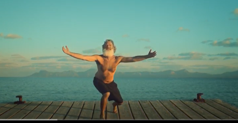 Iggy Pop stands arms wide on a dock with blue sky and ocean in background