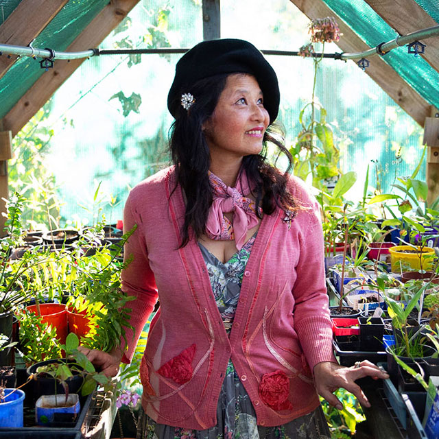 Woman standing in a green house surrounded by plants wearing a pink cardigan 