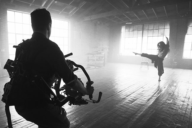 Black and white image of a dancer in a warehouse being videoed by a man holding a video camera