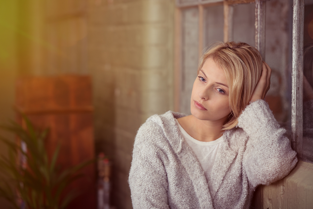 Sad young woman sitting thinking resting her head on her hand on the back of the sofa as she stares at the ground with a serious expression