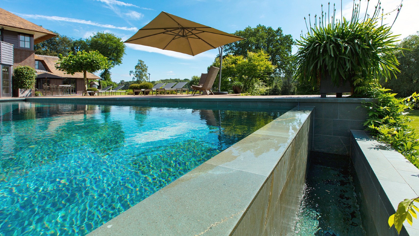 Bouwkundig overloopzwembad met overloop aan het einde in groene tuin met groot huis en parasol