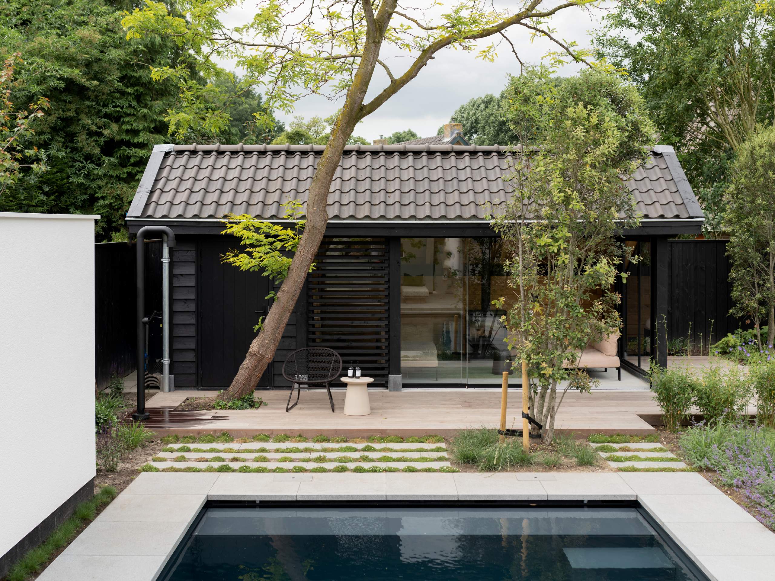 Landelijk poolhouse met pannendak en donkergrijs zwembad ervoor in groene tuin met houten vlonder