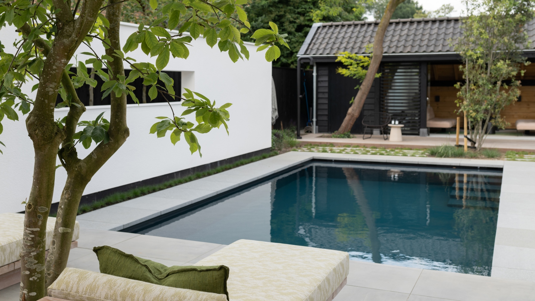 Skimmerzwembad van starline in kleur donkergrijs met lichte randtegels vooraan een ligbed groene boom en achter het zwembad een ruim poolhouse met buitendouche